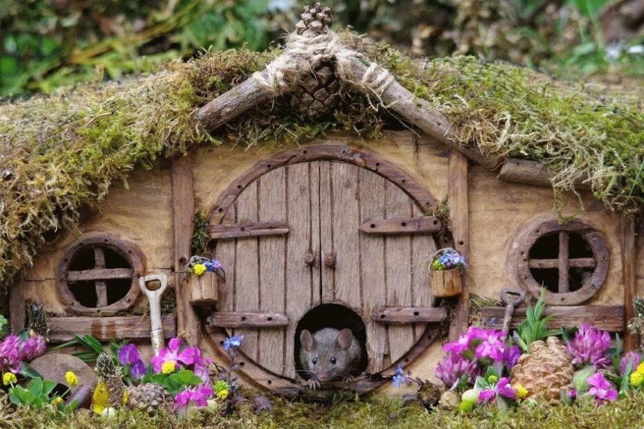 A small mouse peeks out of the round door of a miniature, moss-covered house surrounded by flowers, pinecones, and tiny gardening tools.