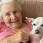 An elderly woman with short white hair sits on a pink armchair, smiling and holding a small light-colored dog in her lap.