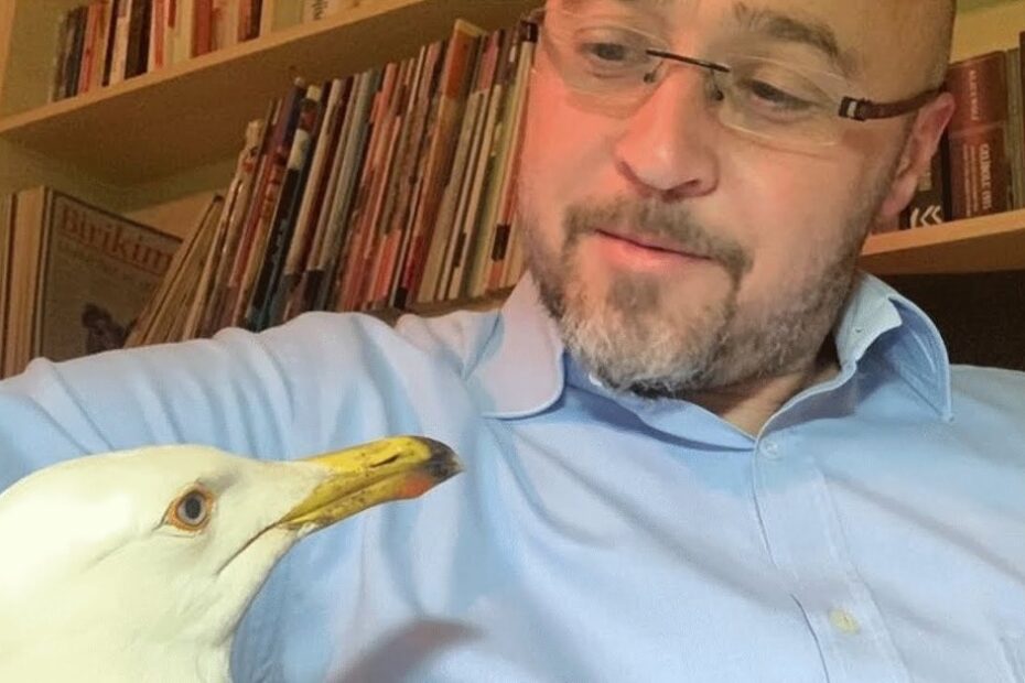 A man with glasses and a beard, wearing a light blue shirt, looks at a seagull perched close to him in a room with shelves of books in the background.