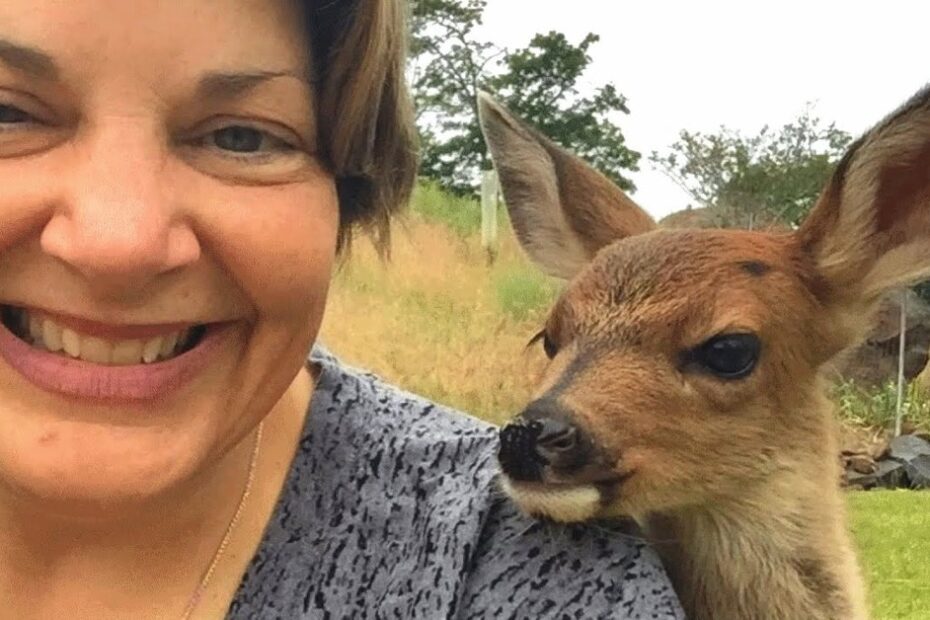 A woman smiles for a selfie outdoors while a young deer stands close beside her, both facing the camera.