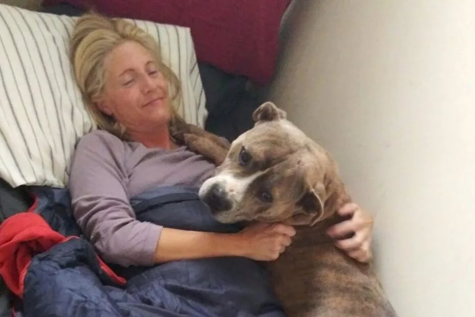A woman lies in bed, smiling with her eyes closed, while hugging a brown and white dog that is resting on her chest.